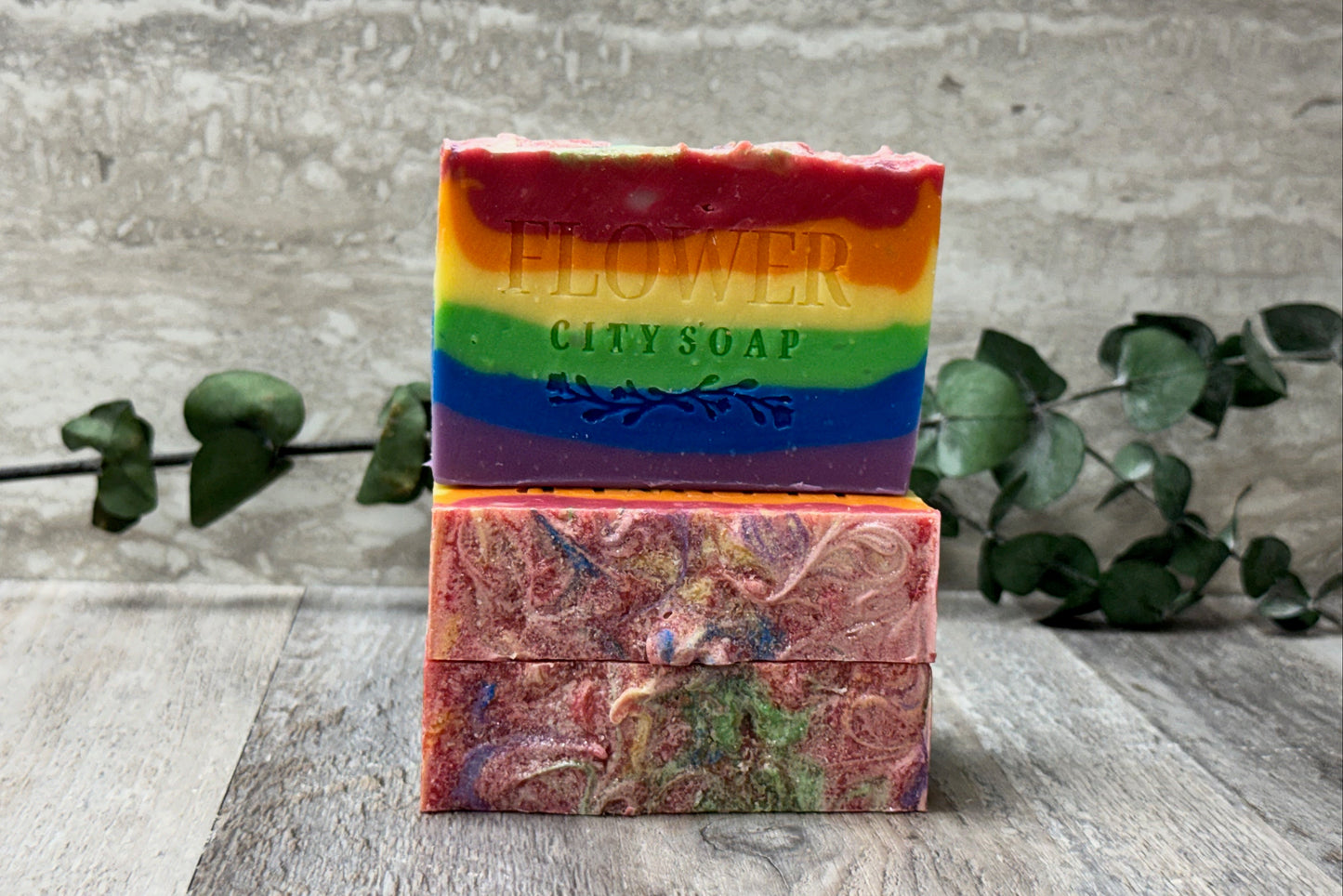 Three handmade soap bars stacked on a rustic wooden surface, with a textured gray wall and green eucalyptus branches in the background. The top bar is a vibrant rainbow-striped soap with the words 'FLOWER CITY SOAP' embossed on the front.