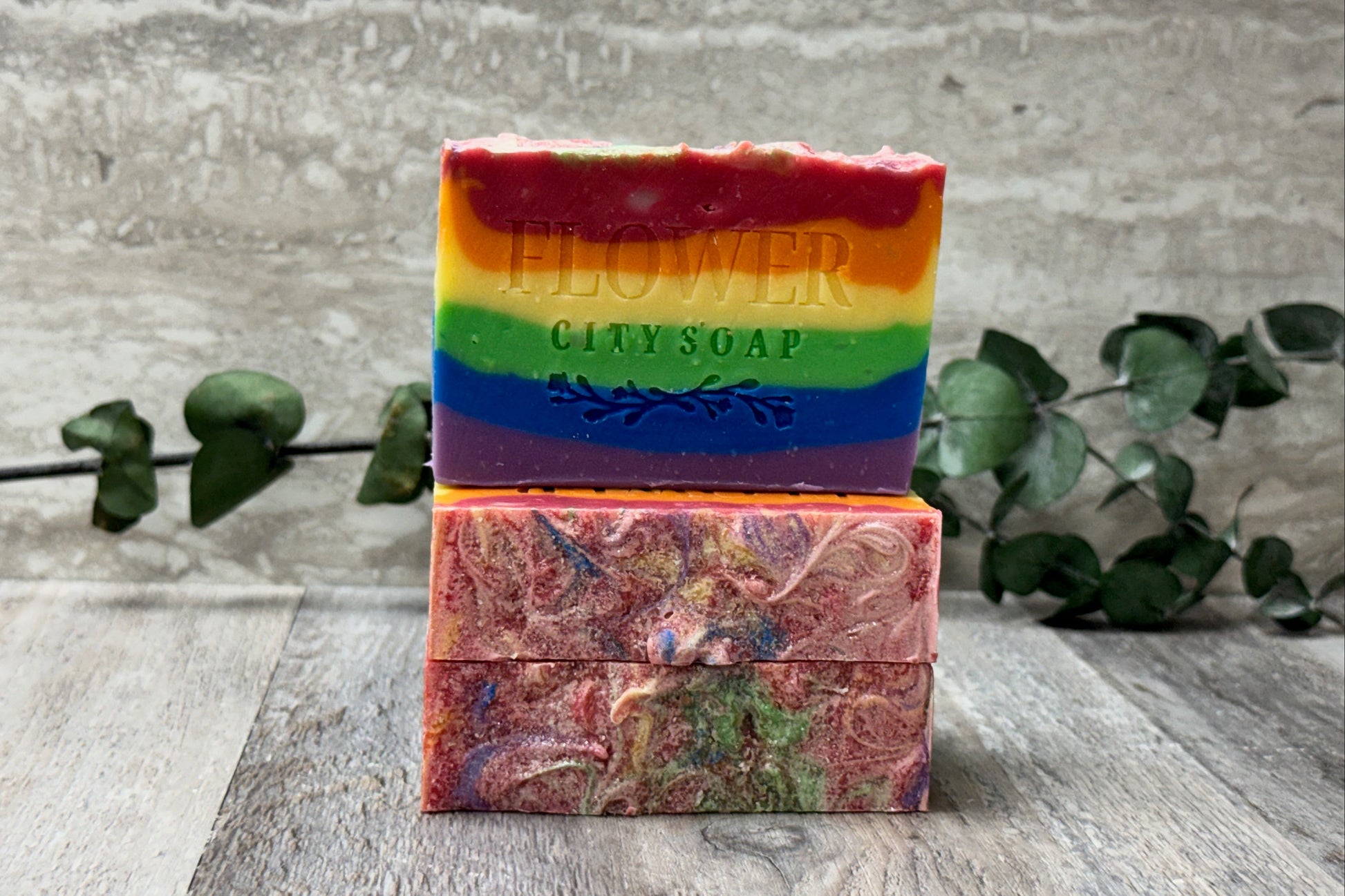 Three handmade soap bars stacked on a rustic wooden surface, with a textured gray wall and green eucalyptus branches in the background. The top bar is a vibrant rainbow-striped soap with the words 'FLOWER CITY SOAP' embossed on the front.