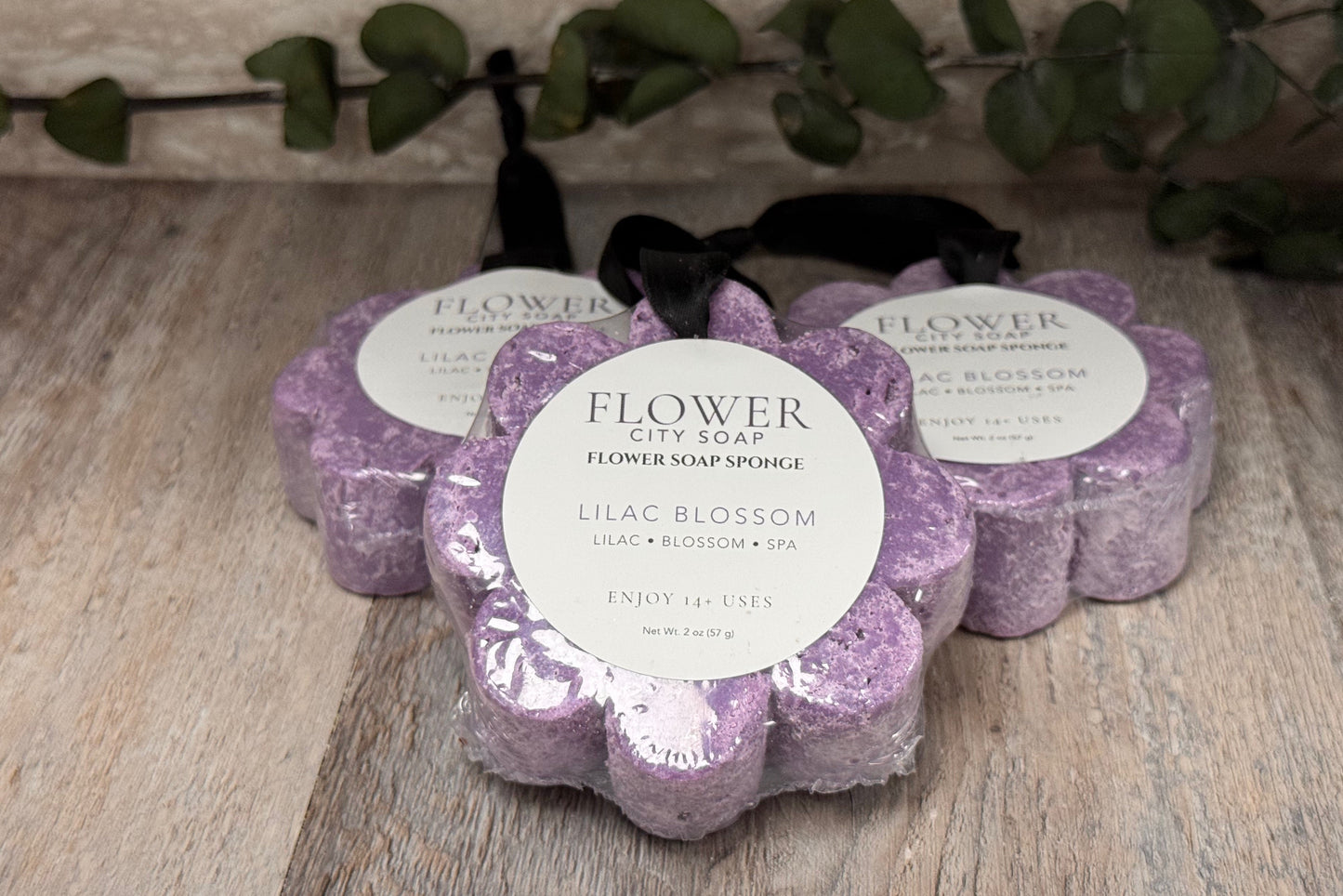 Three flower-shaped flower soap sponges in a Lilac Blossom scent, arranged on a rustic wooden surface with a green leafy vine in the background. Each sponge is wrapped in clear plastic with a white circular label and a black ribbon.