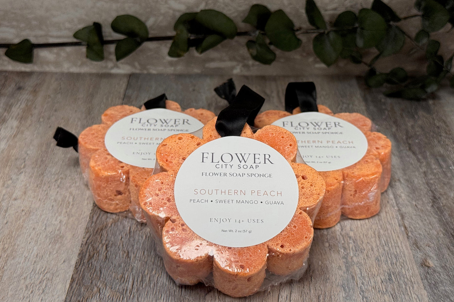 Three flower-shaped flower soap sponges in a Southern Peach scent, arranged on a rustic wooden surface with a green leafy vine in the background. Each sponge is wrapped in clear plastic with a white circular label and a black ribbon.
