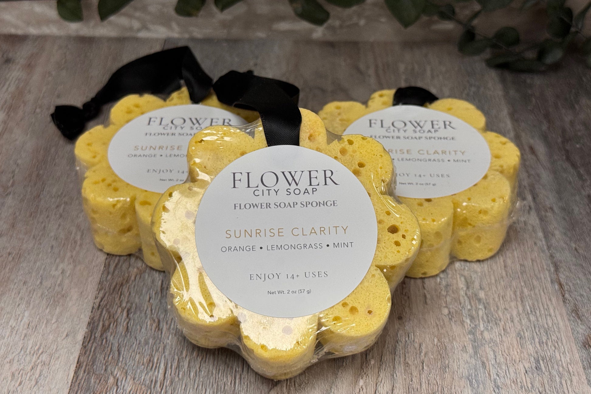 Three yellow flower-shaped flower soap sponges by Flower City Soap in the 'Sunrise Clarity' scent, packaged in clear plastic with white circular labels and black ribbons, arranged on a rustic wooden surface with green leaves in the background.