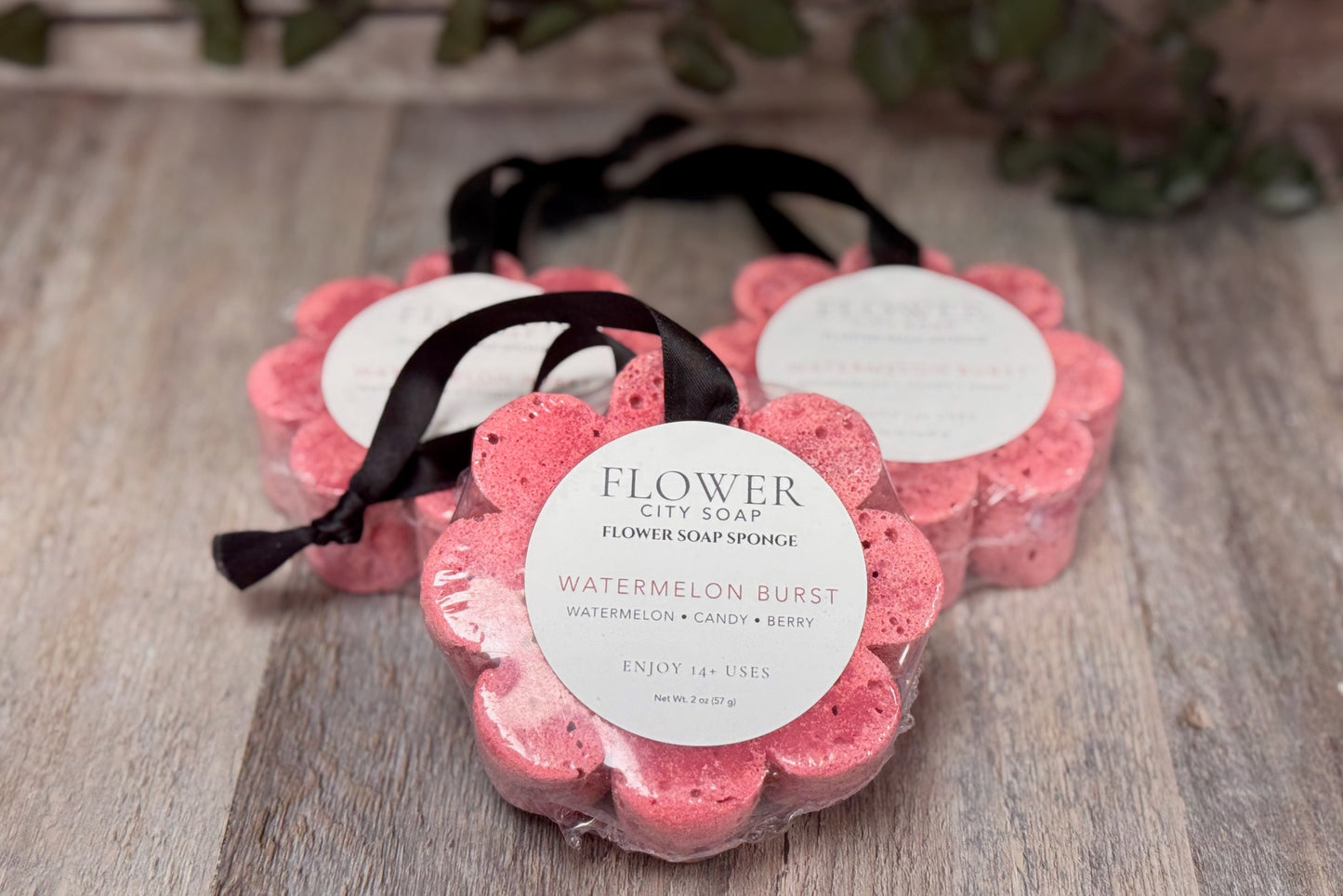 Three flower-shaped flower soap sponges in a Watermelon Burst scent, arranged on a rustic wooden surface with a green leafy vine in the background. Each sponge is wrapped in clear plastic with a white circular label and a black ribbon.