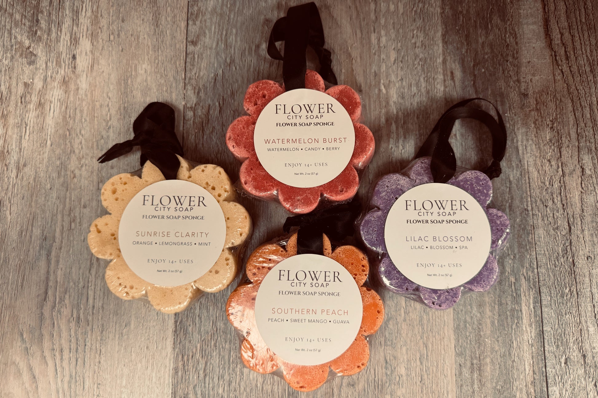 Four flower-shaped flower soap sponges by Flower City Soap arranged on a weathered wood background. The round labels show scents: Sunrise Clarity, Watermelon Splash, Southern Peach, and Lilac Blossom, with black hanging ribbons.