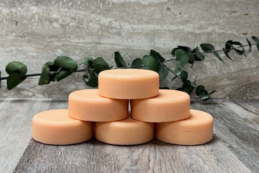 A stack of six round peach-colored bath solid conditioning bars arranged in a pyramid on a gray wood-textured surface, with a green eucalyptus branch behind them against a matching gray stone-look backdrop.
