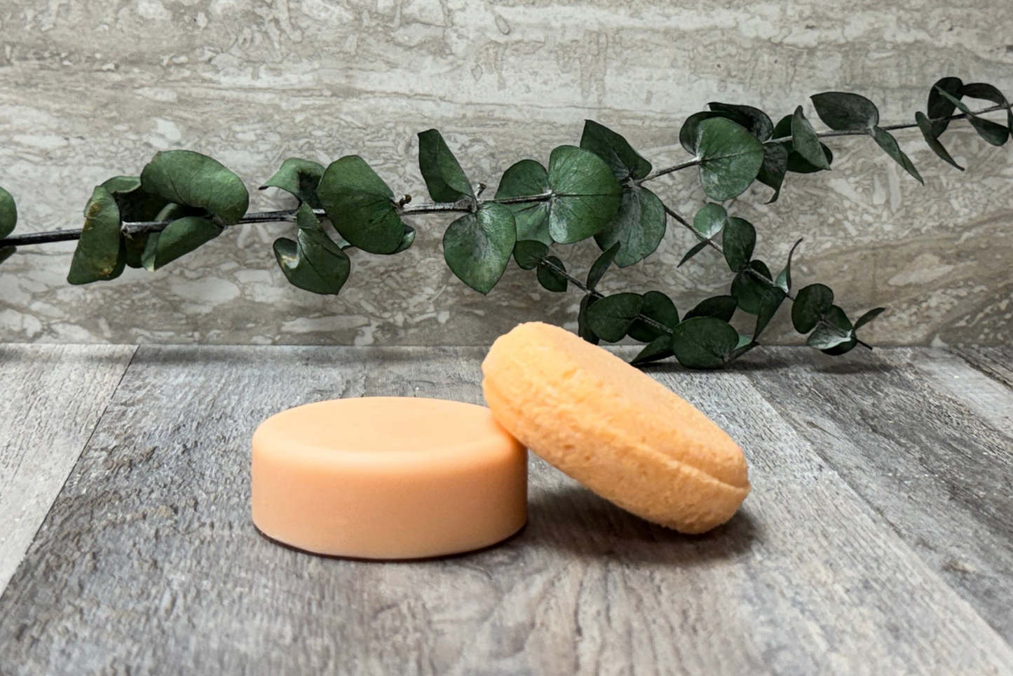 Two round solid shampoo and conditioning bars in peach and light orange-yellow tones, displayed on a gray wood-textured surface with a green leafy branch in the background.